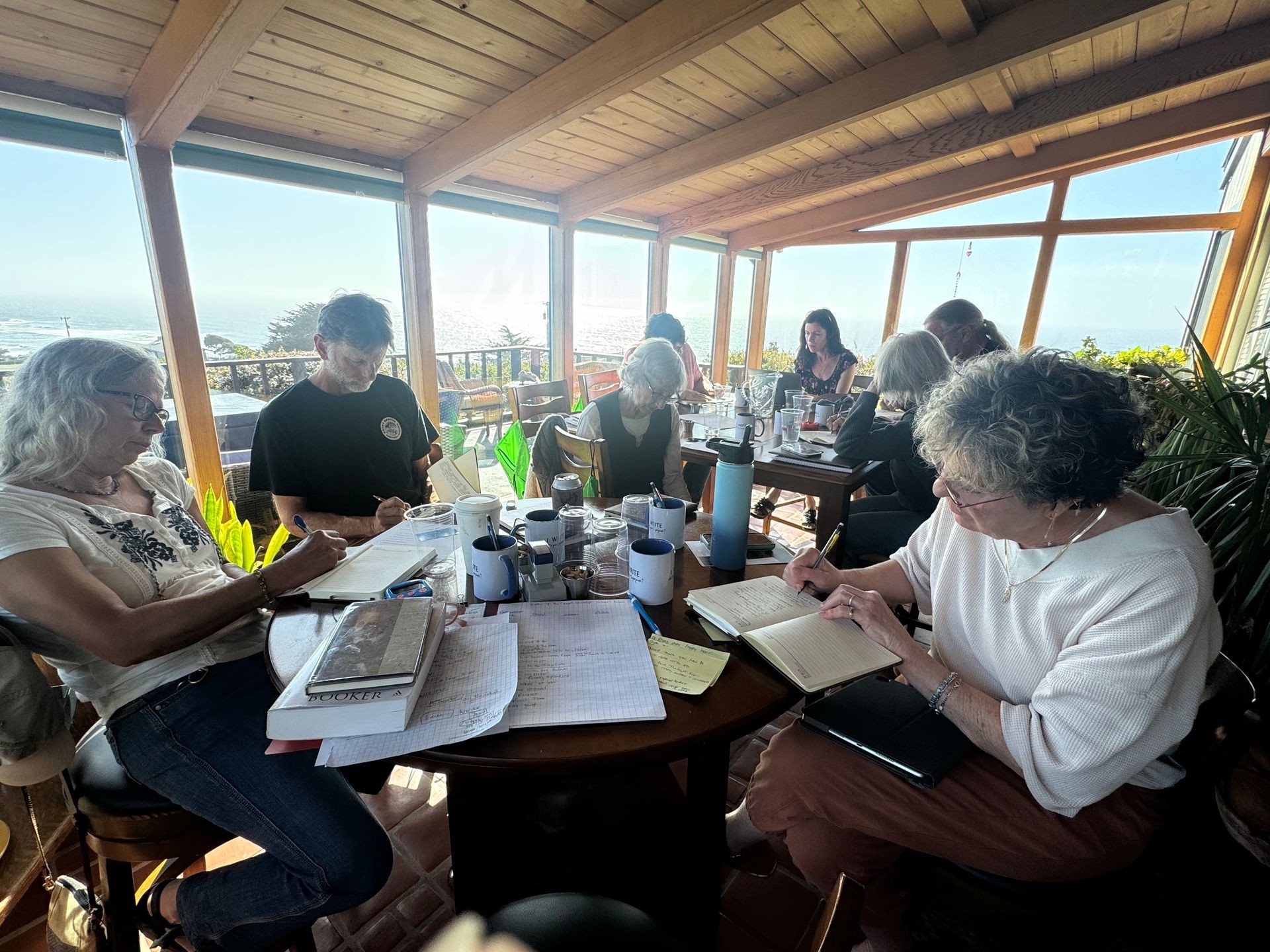 Group of people writing in journals while sitting indoors by large windows overlooking a scenic ocean view.