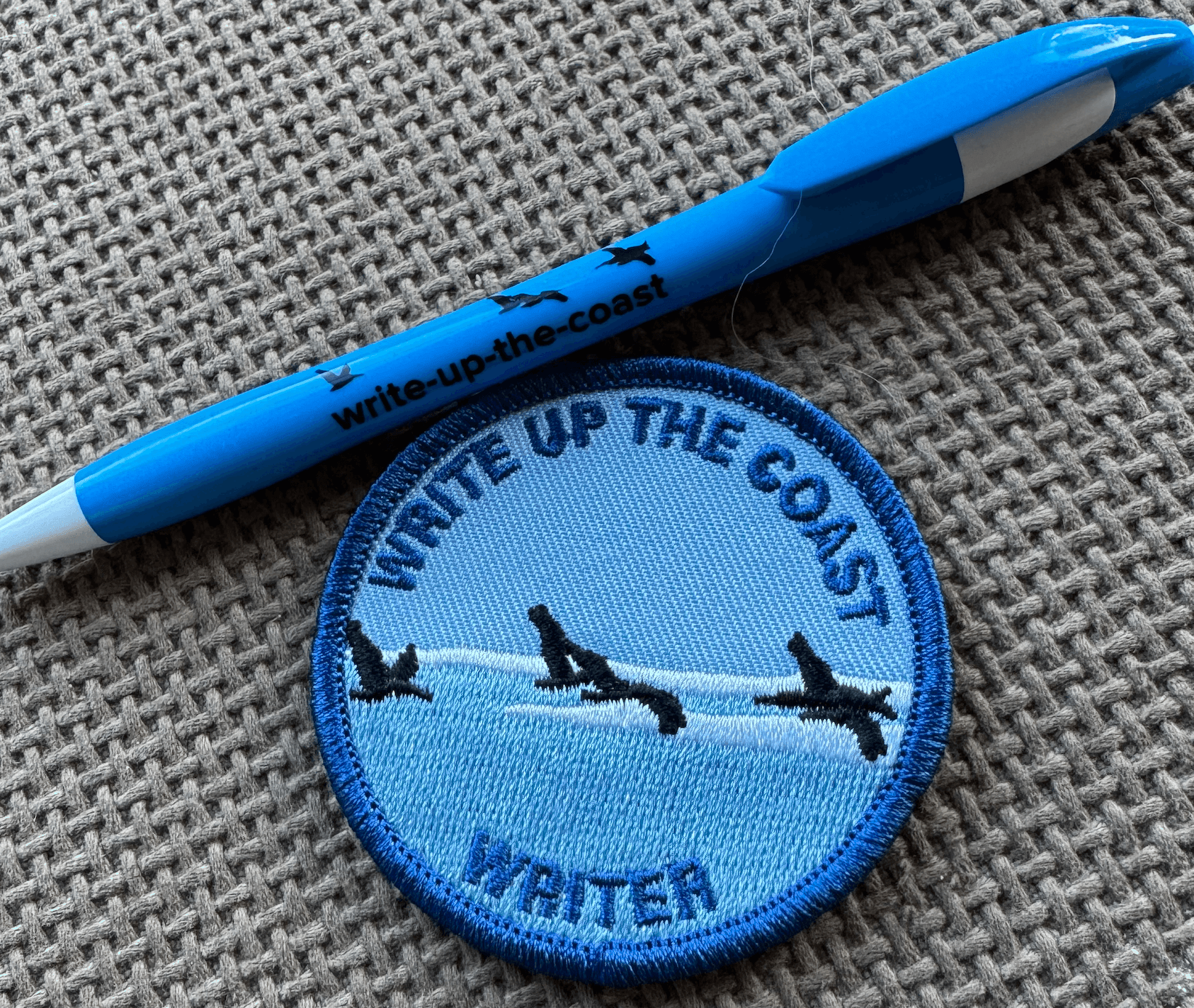 Blue pen and embroidered patch with 'Write Up the Coast' text on textured surface.