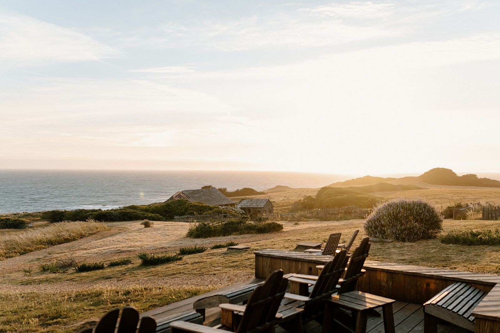 Scenic coastal landscape during sunset with wooden chairs on a deck overlooking the ocean and grassy fields.