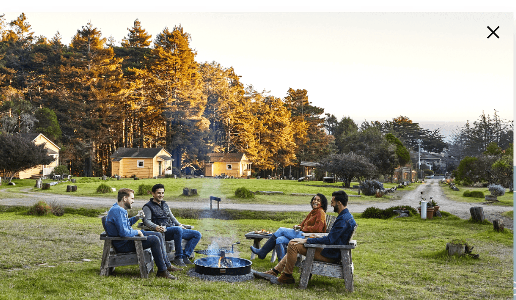 Group of people sitting around a campfire with log cabins and trees in the background at sunset.