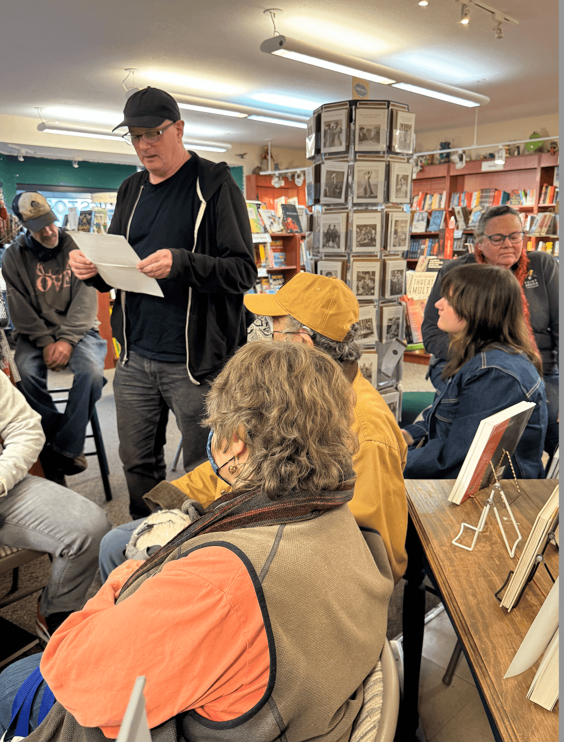 Man in a black jacket reading to a gathered group in a bookstore with shelves and display racks.