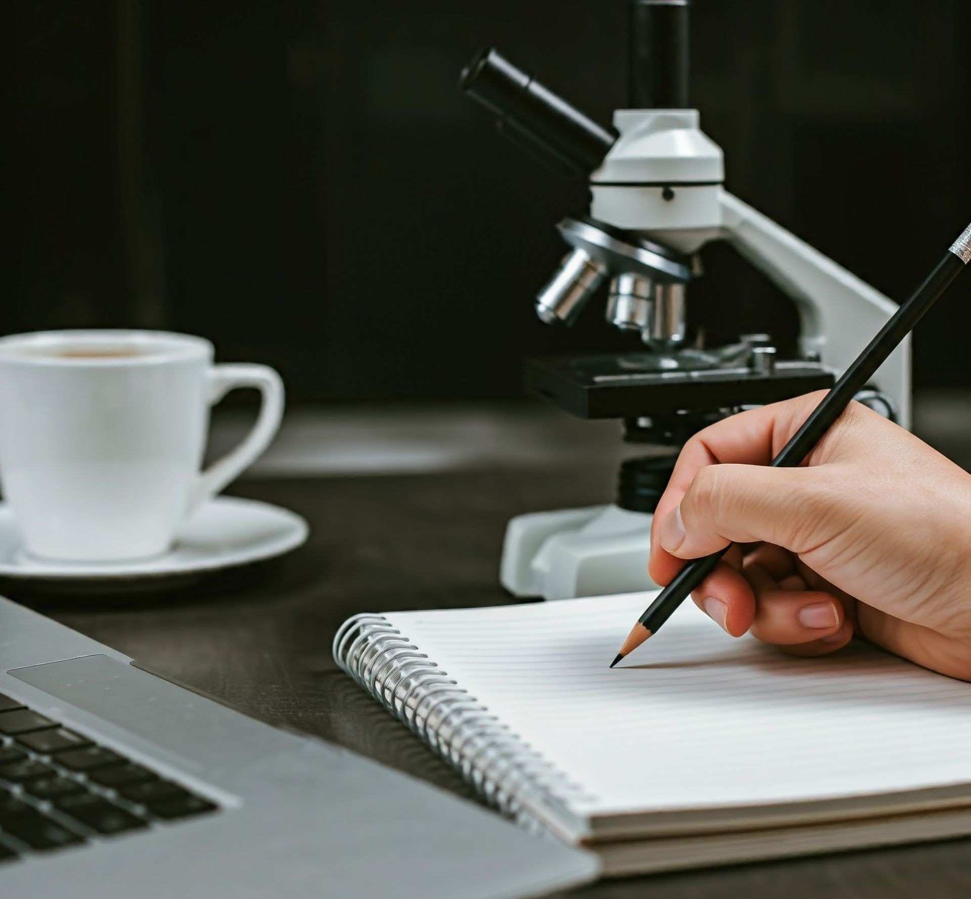 Person writing in a notebook with a pencil beside a microscope, cup of coffee, and laptop.