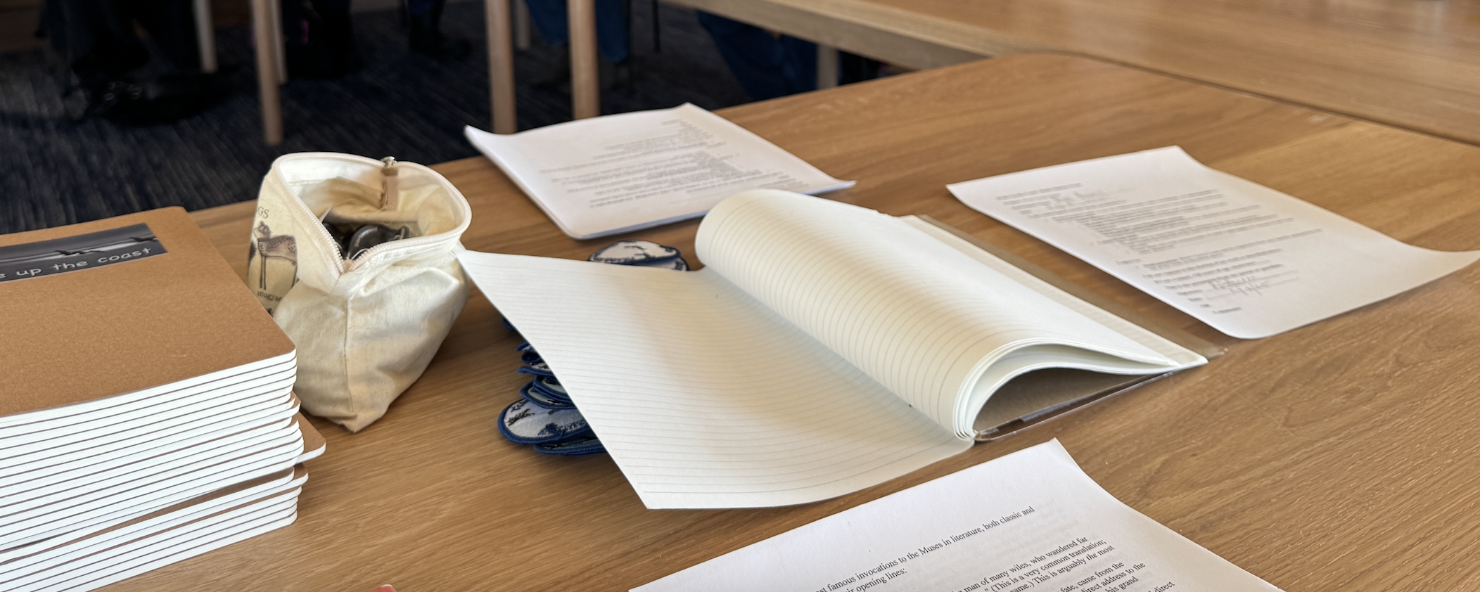 Notebooks, buttons, and pens spread across a wooden table at a Write Up The Coast gathering